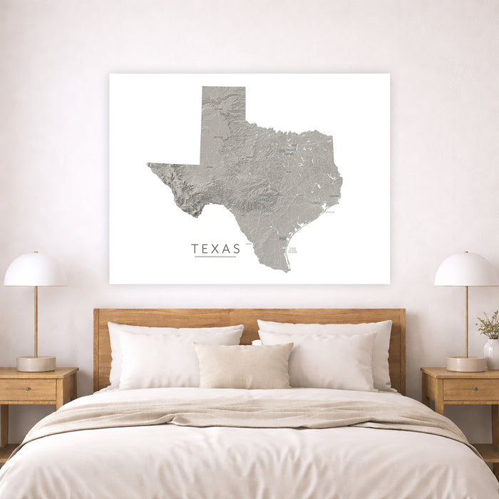 A large white canvas wall art print featuring a gray topographic map of Texas with the word 'TEXAS' below, displayed above a neatly made bed in a modern bedroom setting.