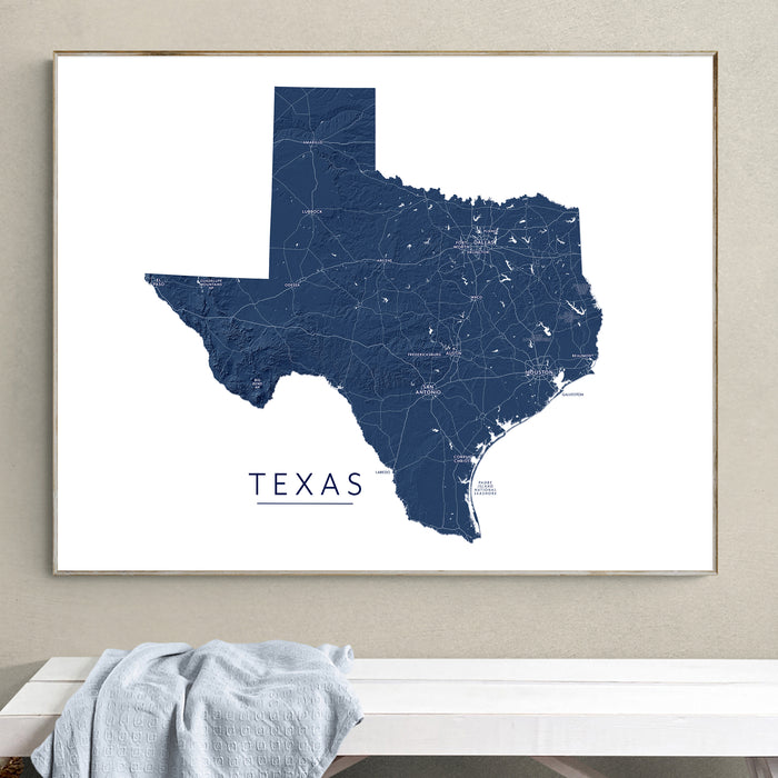 A framed Texas state map print in dark navy blue on a white background, hanging on a beige wall above a white bench with a light blue blanket.