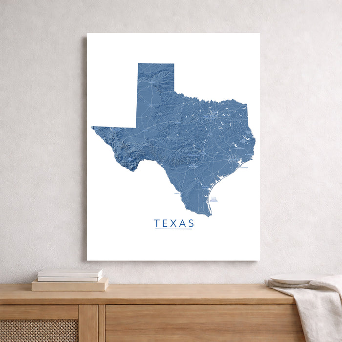 A vertical canvas print featuring a blue topographic map of Texas with the word 'TEXAS' underneath, displayed on a light gray wall above a wooden console table.