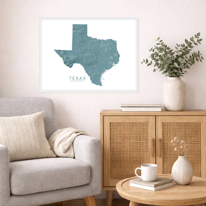 A framed wall art print showing a teal textured map of Texas with the word 'TEXAS' below it, displayed above a light gray armchair and a wooden cabinet in a styled living room.