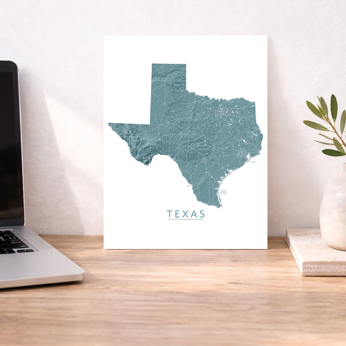 A vertical wall art print featuring a teal topographic map of Texas with the word 'TEXAS' underneath, displayed on a wooden desk next to a laptop and a small plant in a vase.
