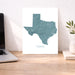 A vertical wall art print featuring a teal topographic map of Texas with the word 'TEXAS' underneath, displayed on a wooden desk next to a laptop and a small plant in a vase.
