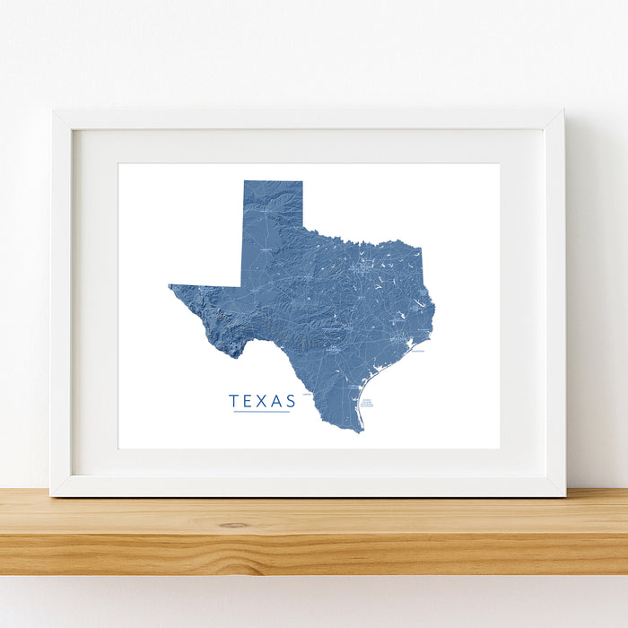 A framed map print of Texas in textured blue tones, displayed on a light wood shelf against a white wall.