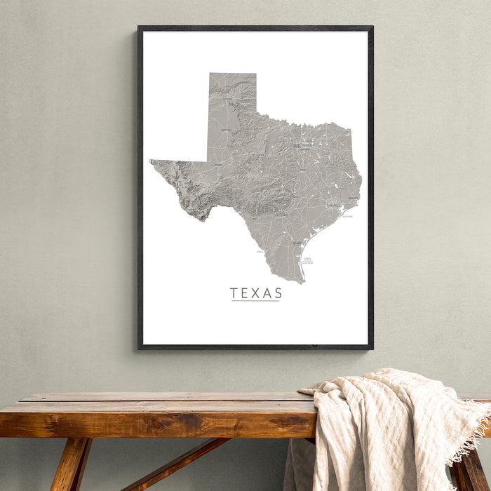 A framed gray map print of Texas hangs on a light gray wall above a wooden bench with a draped cream throw blanket.