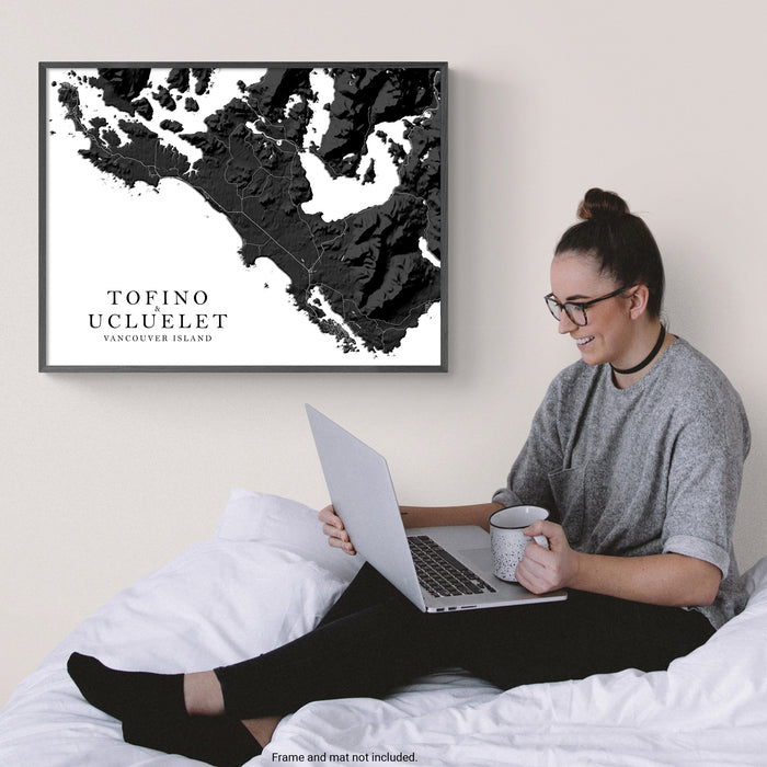 Woman sitting on a bed using a laptop with a framed black and white map of Tofino Ucluelet on the wall.