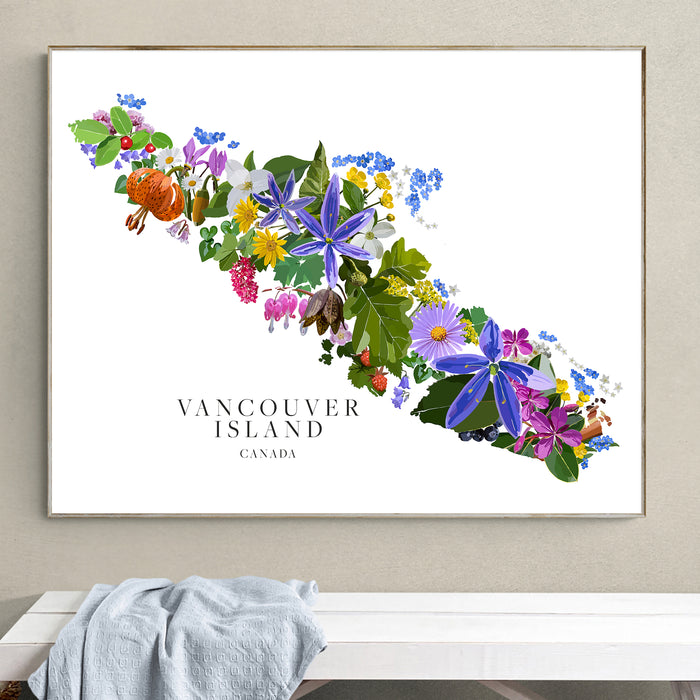 A framed botanical map print of Vancouver Island, Canada, featuring colorful wildflowers arranged along the island's outline on a white background, hung on a beige wall above a white bench with a light blue blanket.