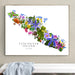 A framed botanical map print of Vancouver Island, Canada, featuring colorful wildflowers arranged along the island's outline on a white background, hung on a beige wall above a white bench with a light blue blanket.