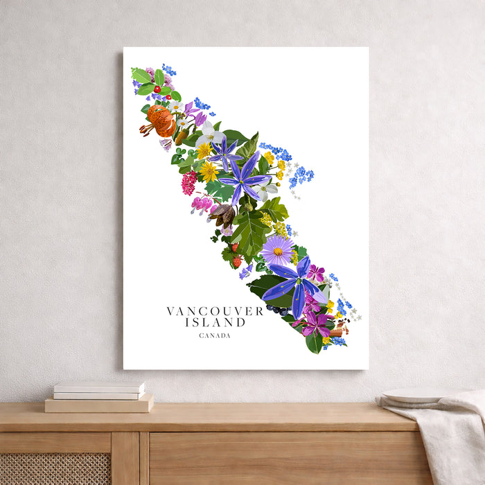 A vertical wall art print featuring a colorful floral map of Vancouver Island, Canada, styled on a light gray wall above a wooden console table with books and a throw blanket.