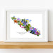A framed watercolor art print of Vancouver Island filled with colorful wildflowers, displayed on a wooden shelf.