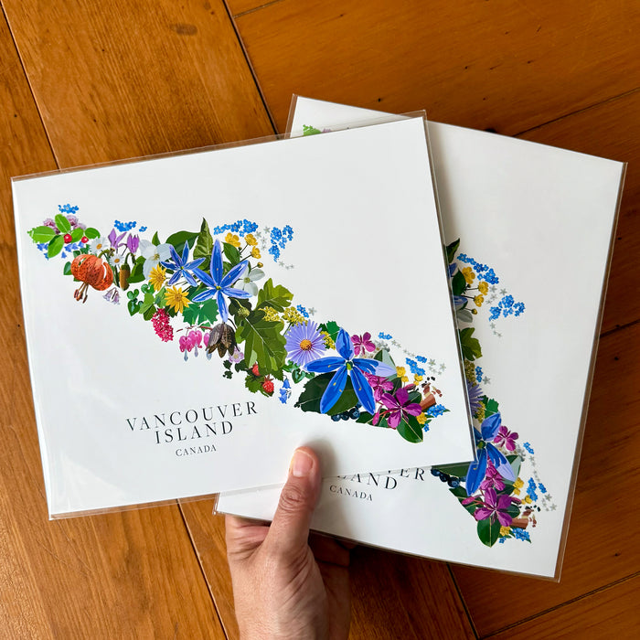 Two square art prints featuring a colorful floral map of Vancouver Island, Canada, held by a hand over a wooden floor background.