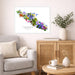 A framed watercolor art print of Vancouver Island filled with colorful wildflowers, displayed on a light beige wall in a styled living room setting with a gray armchair, wooden cabinet, and greenery.