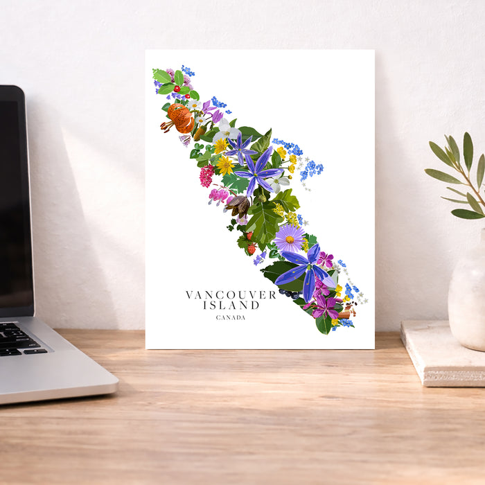 A vertical art print of Vancouver Island decorated with colorful wildflowers and green leaves, displayed on a wooden desk next to a laptop and a small plant in a vase.