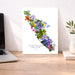 A vertical art print of Vancouver Island decorated with colorful wildflowers and green leaves, displayed on a wooden desk next to a laptop and a small plant in a vase.